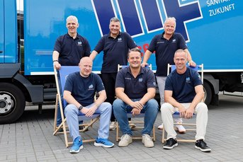 Die Gruppe von Männern steht und sitzt entspannt vor einem großen blauen Fahrzeug, das mit einem Logo versehen ist.