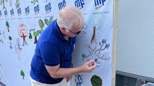 Ein Mann mit blonden Haaren zeichnet an einer Wand mit bunten Baumzeichnungen und Logos im Hintergrund.