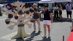 Eine Frau in einem schwarzen T-Shirt und Shorts interagiert mit einem künstlerischen Baum, während ein Mann im Hintergrund zuschaut.
