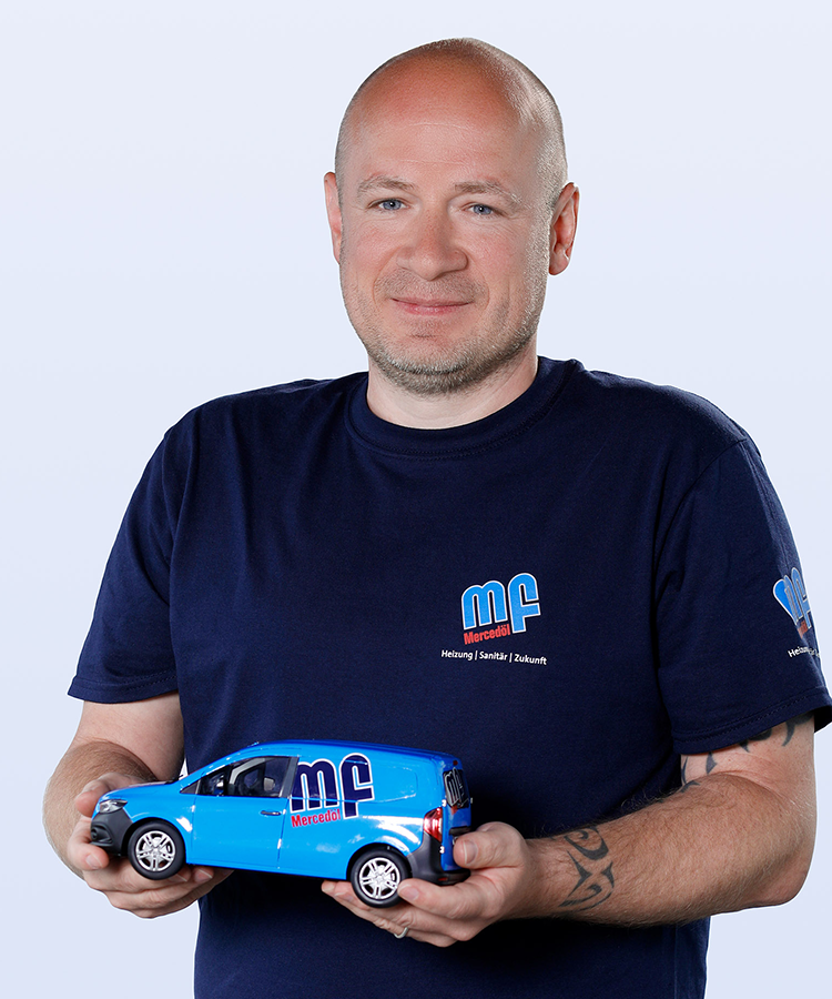 Ein Mann mit Glatze lächelt, während er ein blaues Modellauto mit dem Logo seiner Firma in den Händen hält.