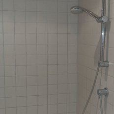 Der Duschbereich zeigt eine saubere, geflieste Wand mit einem Duschregler und einem Seifenspender in Blau.