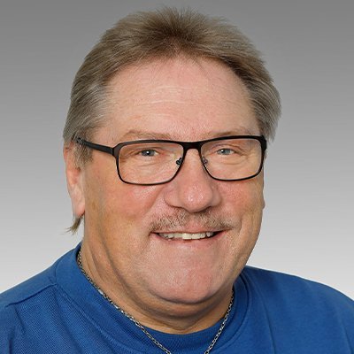 Ein lächelnder Mann mit Brille und einem blauen Pullover posiert vor einem grauen Hintergrund.