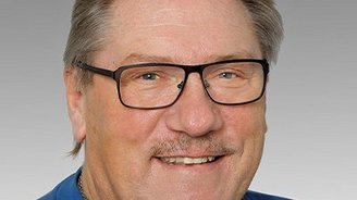 Ein lächelnder Mann mit Brille und einem blauen Pullover posiert vor einem grauen Hintergrund.