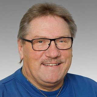 Porträt eines fröhlichen Mannes in blauer Kleidung Ein lächelnder Mann mit Brille und einem blauen Pullover posiert vor einem grauen Hintergrund.