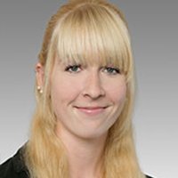 Eine lächelnde Frau mit langen, blonden Haaren und einem professionellen Outfit vor einem grauen Hintergrund.
