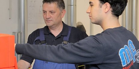 Ein junger Mann zeigt auf ein Gerät, während ein älterer Mann aufmerksam zuschaut, beide in einer Werkstatt mit Maschinen.