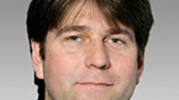 Der Mann hat eine entspannte Haltung und trägt ein schwarzes Hemd, während er direkt in die Kamera schaut.