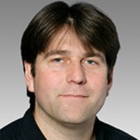 Der Mann hat eine entspannte Haltung und trägt ein schwarzes Hemd, während er direkt in die Kamera schaut.