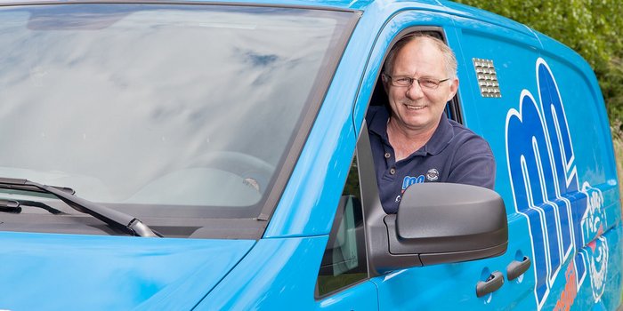 Ein lächelnder Mann sitzt am Steuer eines blauen Lieferwagens und genießt die frische Luft an einem sonnigen Tag.