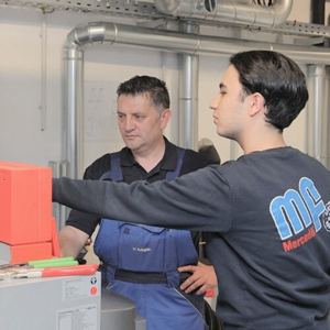 Zwei Männer in Arbeitskleidung stehen in einer Werkstatt, einer bedient ein Gerät, während der andere zuschaut.