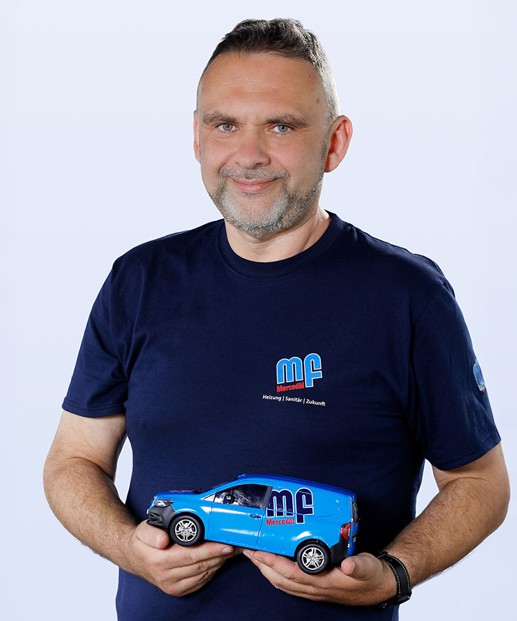 Ein Mann mit einem grauen Bart lächelt, während er ein blaues Modellauto in seinen Händen hält, vor einem hellen Hintergrund.