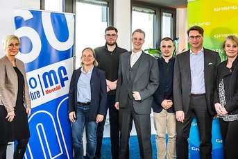 Eine Gruppe von neun Personen steht in einem modernen Büro, umgeben von großen Fenstern und bunten Werbebannern.