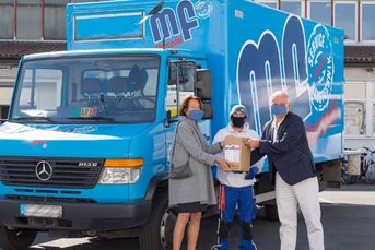 Eine Frau übergibt ein Paket an einen Mann, während ein blauer Lieferwagen im Hintergrund steht, umgeben von weiteren Fahrzeugen.