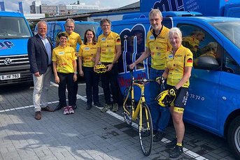 Eine Gruppe von Radfahrern in gelben Trikots steht neben blauen Lieferwagen, bereit für ein bevorstehendes Event.