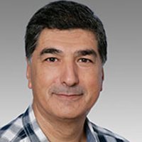 Ein Mann mit dunklen Haaren und einem freundlichen Lächeln trägt ein kariertes Hemd und schaut direkt in die Kamera.
